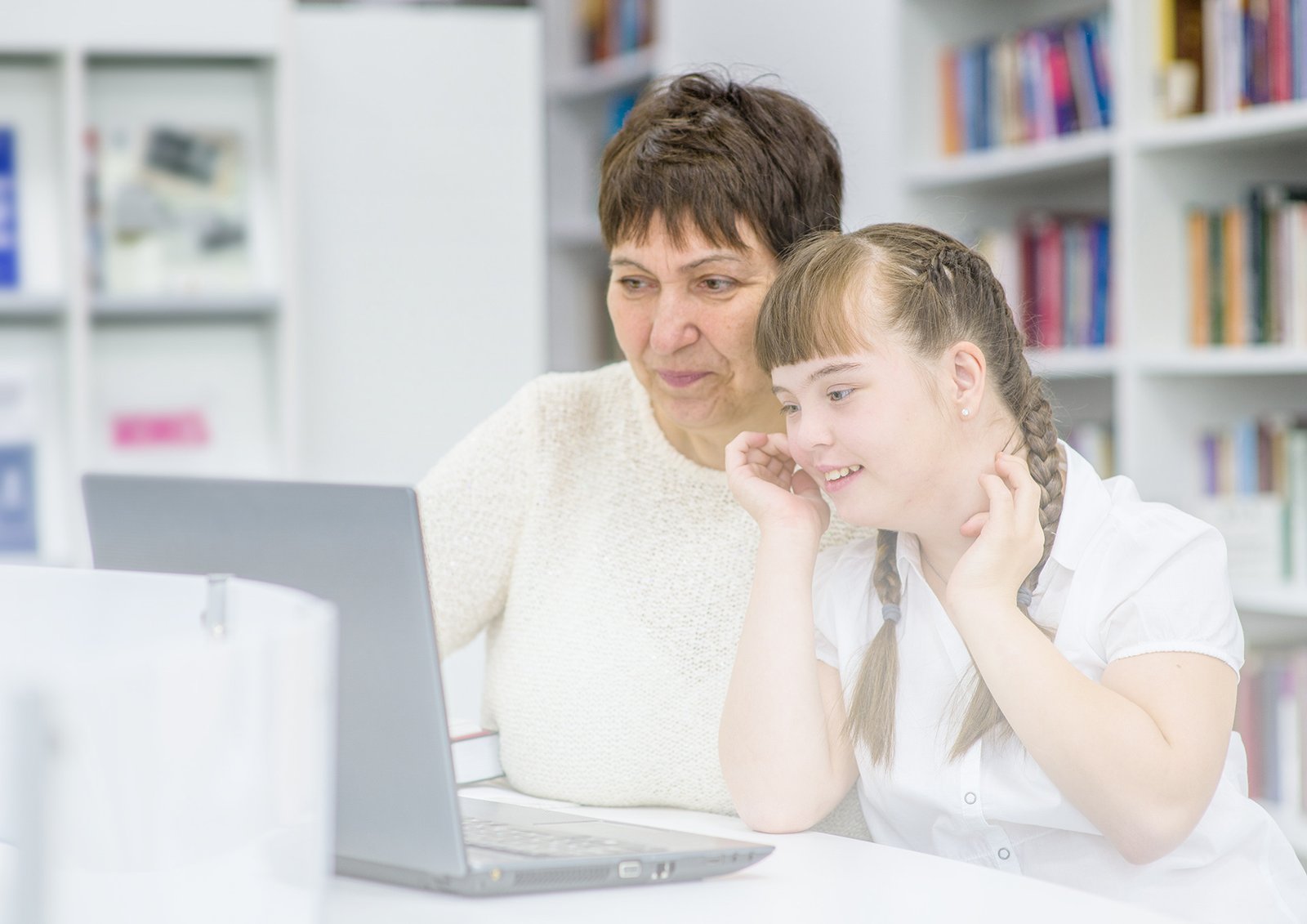 An older woman teaching a delighted teenage student with Down Syndrome. They are both looking at a laptop.