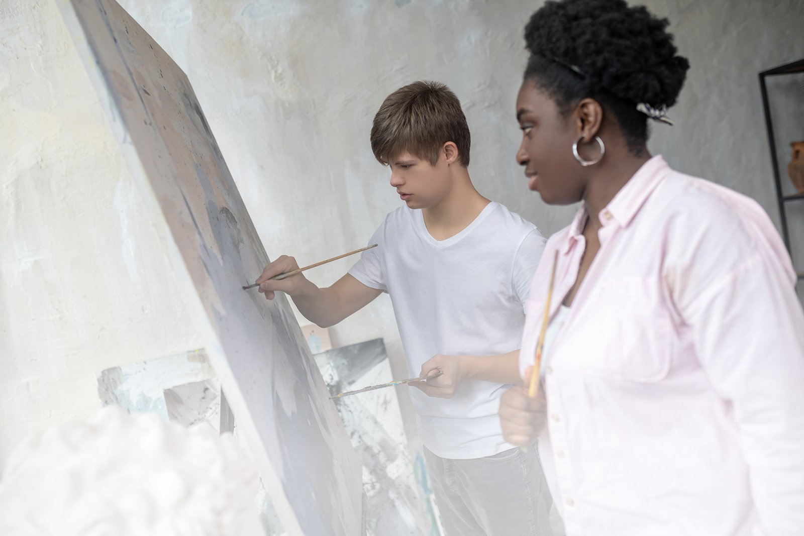 A young teacher guides a teenage student with Down Syndrome in painting on a canvas.