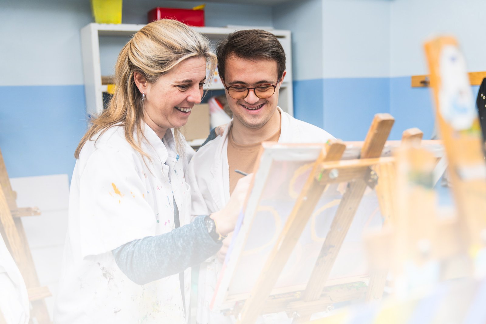 An art teacher enthusiastically demonstrates working on a canvas while a student with Down Syndrome looks on.