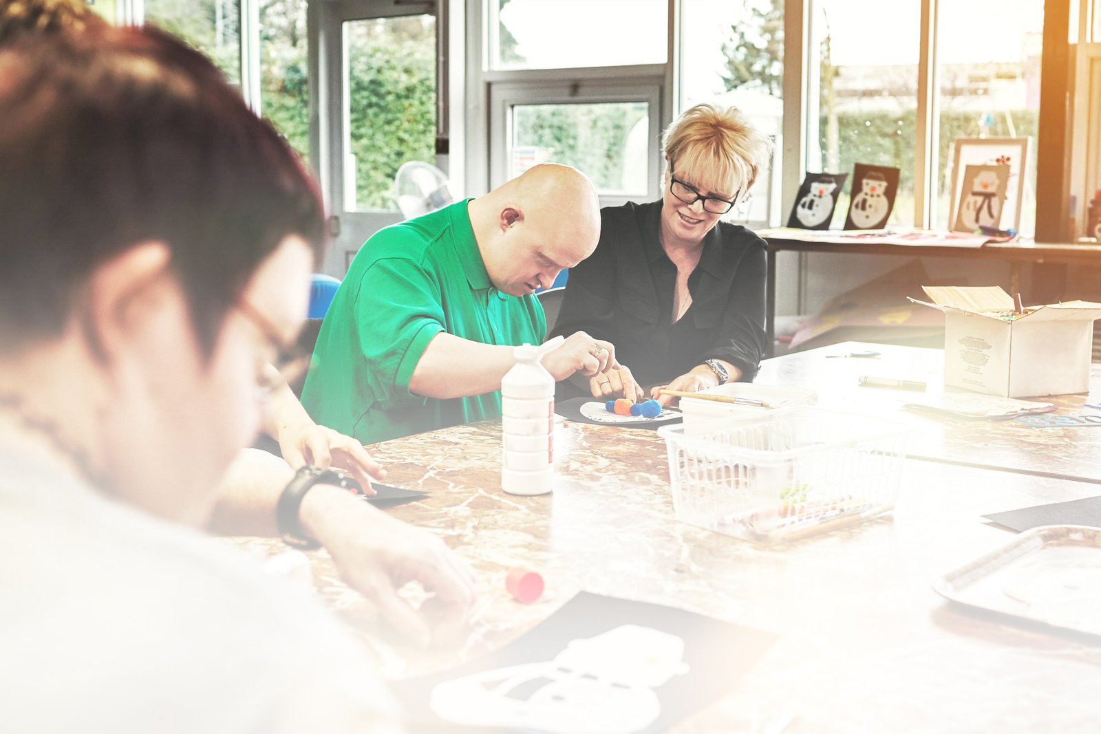 At a large table with several students, a female teacher guides a student with Down Syndrome through an art project.